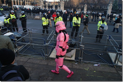 A pink Star Wars Stormtrooper walks past riot police